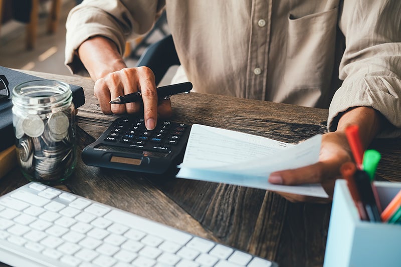 Person using a calculator while reviewing financial documents at a desk, with a keyboard, jar of coins, and office supplies visible in the workspace
